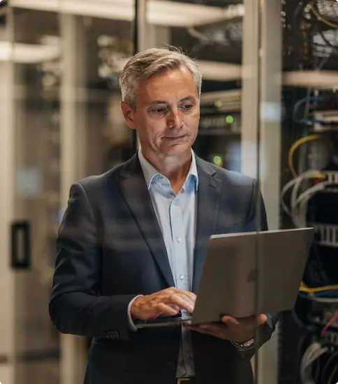 A man in his 50s stood in a server room while working on a laptop