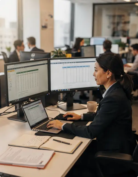 Image of a woman sat in a busy modern office. She is working at a laptop with multiple screens